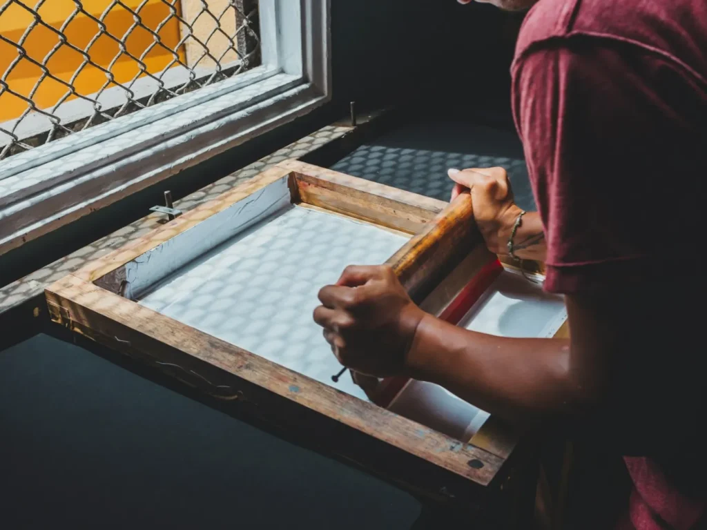 Homem segura quadro de serigrafia junto à janela, estampagem por DTF, serigrafia e sublimação