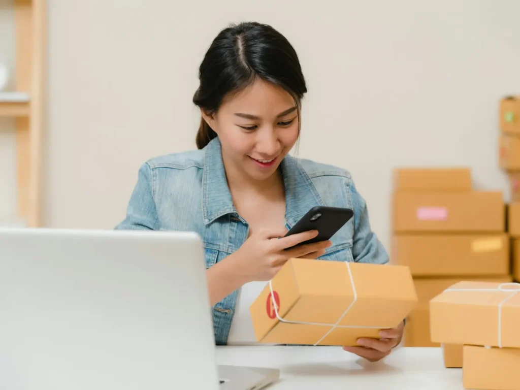 Mulher a verificar encomenda no telemóvel junto a caixas, Google Shopping para lojas online